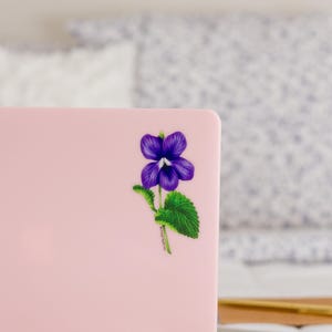 May include: A pink laptop computer with a purple flower sticker on the lid. The flower has green leaves and a white center.