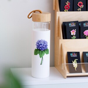Puede incluir: Una botella de agua de vidrio con tapa de bamb&uacute; y una correa marr&oacute;n. La botella tiene un fondo blanco con una ilustraci&oacute;n de flor de hortensia morada y el texto "Hydrangea".