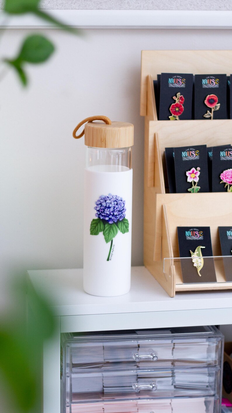 Puede incluir: Una botella de agua de vidrio con tapa de bamb&uacute; y una correa marr&oacute;n. La botella tiene un fondo blanco con una ilustraci&oacute;n de flor de hortensia morada y el texto "Hydrangea".