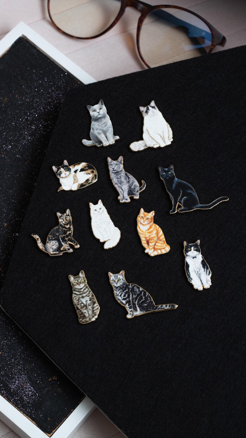 May include: A collection of 12 enamel pins featuring different cat breeds. The pins are all in various colors and poses, including a black cat, a tabby cat, a calico cat, and a white cat.