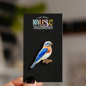 May include: A bluebird enamel pin with gold-tone metal edges, featuring a blue head, wings, and tail with a rust-colored chest and white belly. The pin is attached to a black rectangular display card with the text THE GRAY MUSE printed in white.