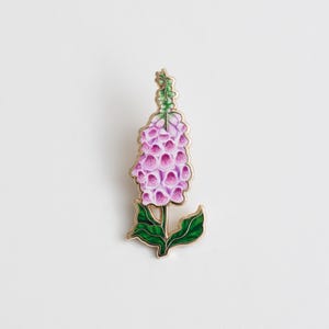 May include: A gold-tone enamel pin featuring a pink and green foxglove flower.