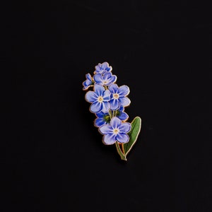 May include: A gold-toned enamel pin featuring a cluster of forget-me-not flowers with blue petals and green leaves.