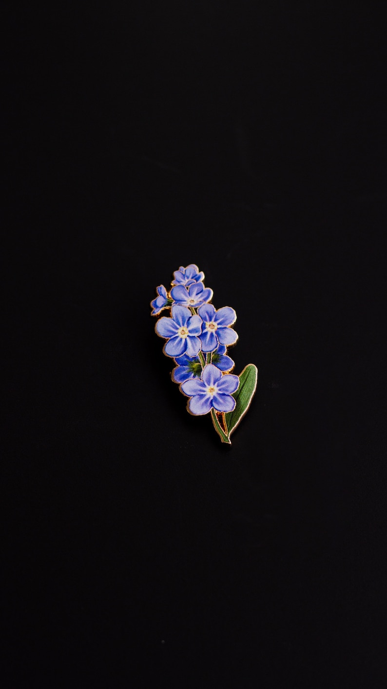 May include: A gold-toned enamel pin featuring a cluster of forget-me-not flowers with blue petals and green leaves.