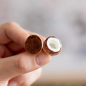 May include: Gold-toned coconut earrings. One earring shows the exterior of the coconut, with brown textured detail. The other earring shows the interior, with white and beige details. The earrings are round and have a gold border.