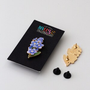 May include: A gold-colored enamel pin featuring a cluster of blue forget-me-not flowers with green leaves. The pin is on a black card with the text "The Gray Muse".