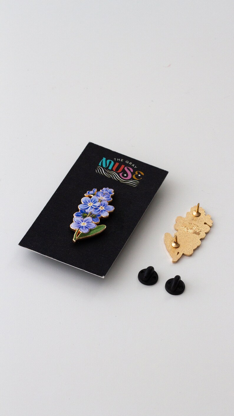 May include: A gold-colored enamel pin featuring a cluster of blue forget-me-not flowers with green leaves. The pin is on a black card with the text "The Gray Muse".