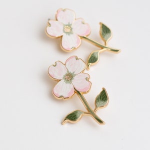 K&ouml;nnte beinhalten: Zwei bl&uuml;tenf&ouml;rmige Broschen. Jede Brosche hat eine rosa-wei&szlig;e Blume mit goldfarbenem Umriss und Stiel. Die Stiele haben gr&uuml;ne Bl&auml;tter. Die Broschen sind vor einem wei&szlig;en Hintergrund platziert.