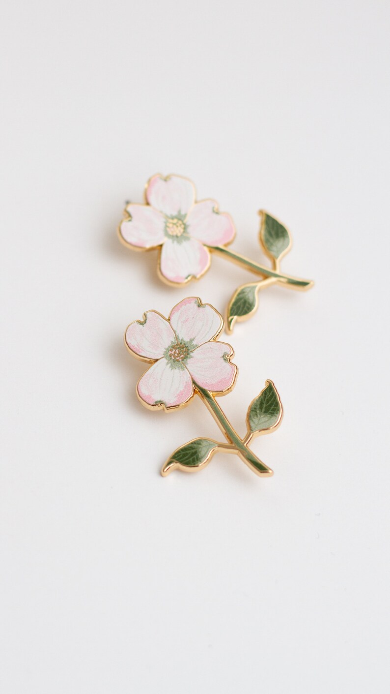 K&ouml;nnte beinhalten: Zwei bl&uuml;tenf&ouml;rmige Broschen. Jede Brosche hat eine rosa-wei&szlig;e Blume mit goldfarbenem Umriss und Stiel. Die Stiele haben gr&uuml;ne Bl&auml;tter. Die Broschen sind vor einem wei&szlig;en Hintergrund platziert.