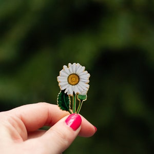 May include: A white daisy with a yellow centre, with green leaves and a bud, is held in a hand with pink nail polish. The background is a blurred green.
