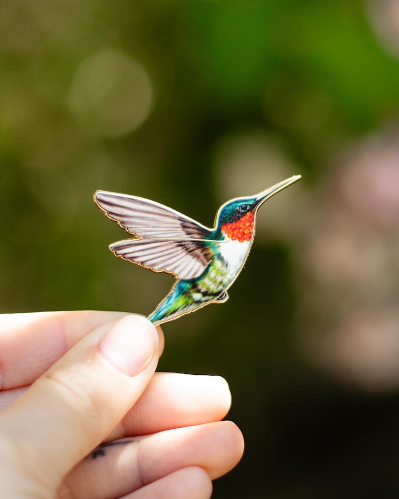 Op de afbeelding: Een kleurrijke kolibrie-broche met een gouden rand. De vogel heeft een rode keel, een blauwe kop, een witte borst en groene en blauwe vleugels en staart. De kolibrie is in volle vlucht afgebeeld. De broche wordt in een hand gehouden.