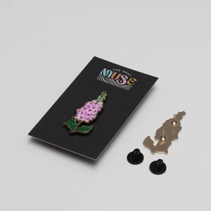May include: A pink and green enamel pin of a flower with the text "The Gray Muse" on a black card.