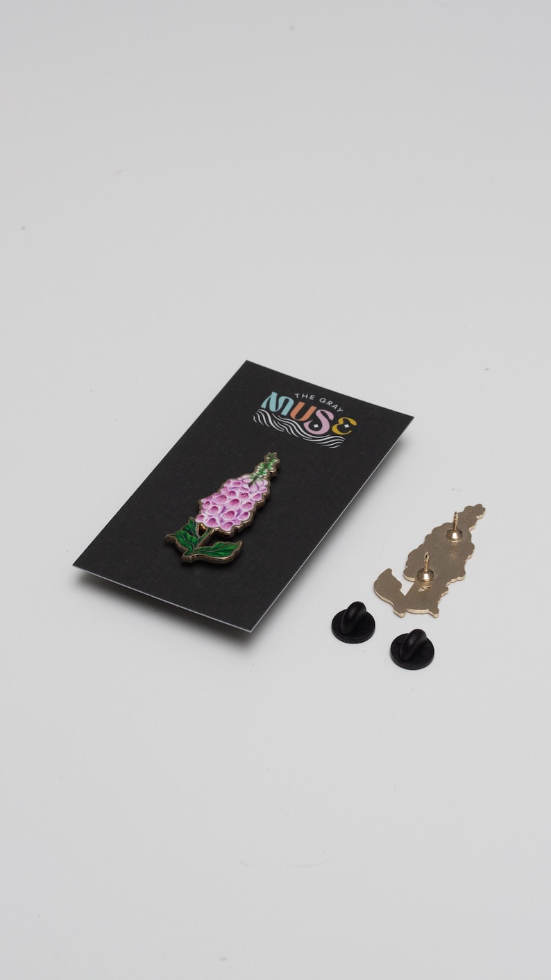 May include: A pink and green enamel pin of a flower with the text "The Gray Muse" on a black card.