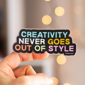 May include: A clear sticker with the text "CREATIVITY NEVER GOES OUT OF STYLE" in colorful letters on a black background. The sticker is being held by a hand against a blurred background of white and gold lights.