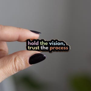 May include: A black enamel pin with a gold border and the text "hold the vision, trust the process" in colorful letters.