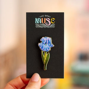 May include: A gold-toned enamel pin featuring a blue iris flower with green leaves. The pin is on a black card with the text "The Gray Muse" in colorful lettering.