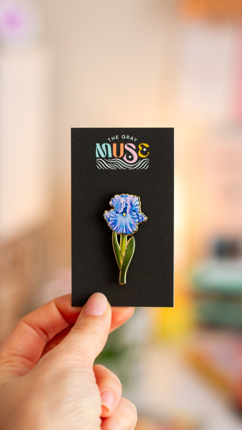 May include: A gold-toned enamel pin featuring a blue iris flower with green leaves. The pin is on a black card with the text "The Gray Muse" in colorful lettering.