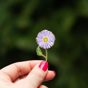Puede incluir: Un pin de flor de esmalte morado con un centro amarillo y hojas verdes. El pin está siendo sostenido por una mano con esmalte de uñas rosa.