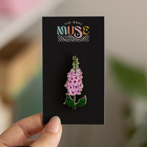 May include: A gold-toned enamel pin featuring a pink and green foxglove flower. The pin is displayed on a black card with the text "The Gray Muse" in colorful lettering.