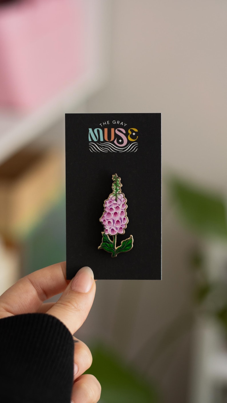 May include: A gold-toned enamel pin featuring a pink and green foxglove flower. The pin is displayed on a black card with the text "The Gray Muse" in colorful lettering.