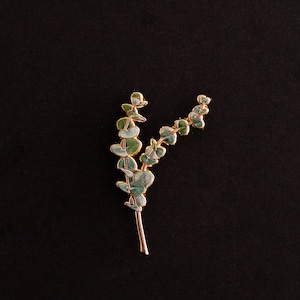 May include: A gold-coloured eucalyptus leaf brooch with green and blue-green enamel leaves. The brooch is set against a black background, highlighting the delicate details of the botanical design. This accessory is suitable for adding a touch of nature-inspired elegance.