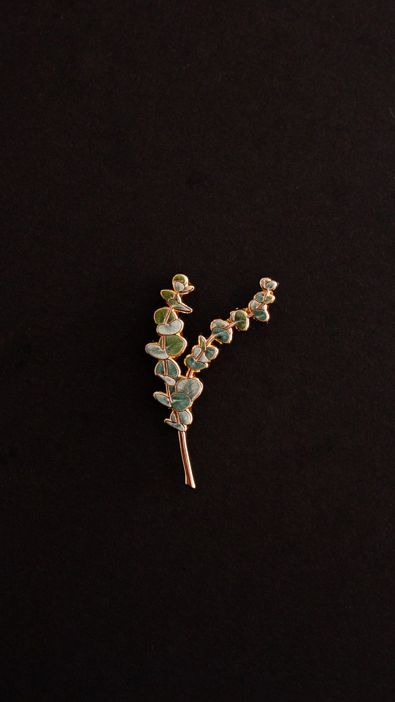 May include: A gold-coloured eucalyptus leaf brooch with green and blue-green enamel leaves. The brooch is set against a black background, highlighting the delicate details of the botanical design. This accessory is suitable for adding a touch of nature-inspired elegance.