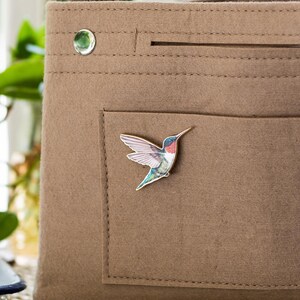 Op de afbeelding: Een bruine vilten tote bag met een klein vakje, een zilveren drukknop en een kolibrie-speld. De speld heeft een kleurrijke kolibrie met blauwe, groene, rode en witte details. De tas heeft een eenvoudig, minimalistisch ontwerp.