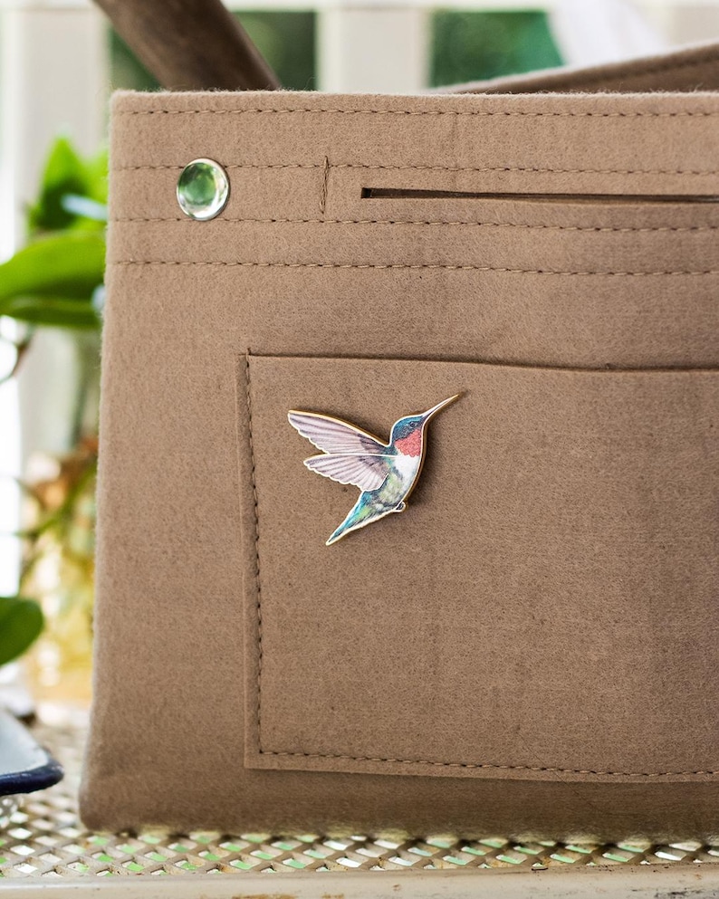 Op de afbeelding: Een bruine vilten tote bag met een klein vakje, een zilveren drukknop en een kolibrie-speld. De speld heeft een kleurrijke kolibrie met blauwe, groene, rode en witte details. De tas heeft een eenvoudig, minimalistisch ontwerp.
