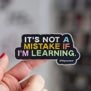 May include: A clear sticker with a black background and colourful text that reads "It's not a mistake if I'm learning." The text is in a rainbow colour scheme. The sticker is held by a finger with pink nail polish.