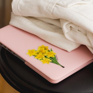 K&ouml;nnte beinhalten: Ein rosa Laptop mit einem gelben Narzissen-Aufkleber auf dem Deckel. Der Laptop steht auf einem schwarzen Tisch.