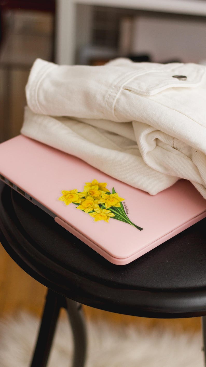 K&ouml;nnte beinhalten: Ein rosa Laptop mit einem gelben Narzissen-Aufkleber auf dem Deckel. Der Laptop steht auf einem schwarzen Tisch.