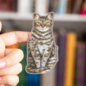 May include: A gray and white tabby cat sticker with yellow eyes. The cat is sitting with its paws tucked under its body. The sticker is on a white background.