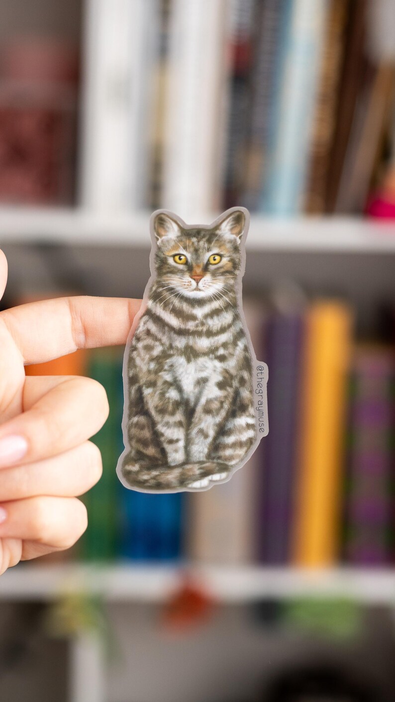 May include: A gray and white tabby cat sticker with yellow eyes. The cat is sitting with its paws tucked under its body. The sticker is on a white background.
