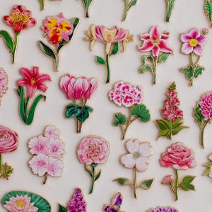 K&ouml;nnte beinhalten: Eine Sammlung farbenfroher Blumen-Emaille-Anstecknadeln mit goldfarbenem Rand. Die Anstecknadeln zeigen verschiedene Blumen, darunter Rosen, Lilien und Sonnenblumen, in Gelb-, Rosa-, Lila- und Rott&ouml;nen. Die Anstecknadeln sind auf wei&szlig;em Hintergrund angeordnet.