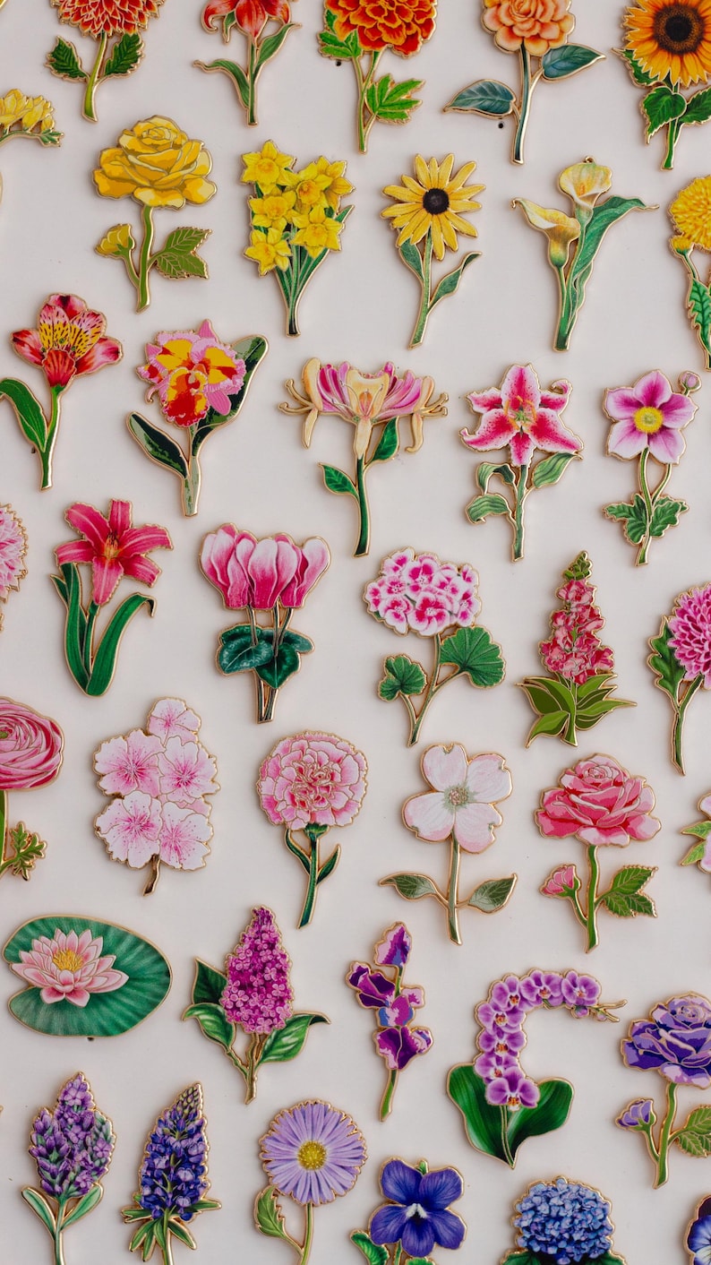 K&ouml;nnte beinhalten: Eine Sammlung farbenfroher Blumen-Emaille-Anstecknadeln mit goldfarbenem Rand. Die Anstecknadeln zeigen verschiedene Blumen, darunter Rosen, Lilien und Sonnenblumen, in Gelb-, Rosa-, Lila- und Rott&ouml;nen. Die Anstecknadeln sind auf wei&szlig;em Hintergrund angeordnet.