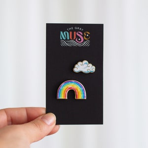 May include: A black card with the text "The Gray Muse" in colorful letters. The card features two enamel pins: a rainbow and a white cloud.