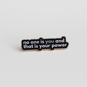 May include: Black enamel pin with gold trim featuring the text "no one is you and that is your power" in white lettering.