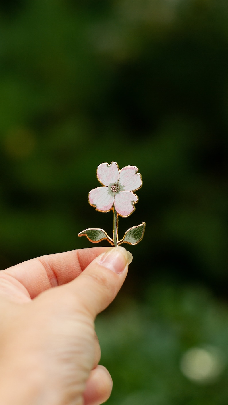 K&ouml;nnte beinhalten: Zarte Blumenbrosche mit einer vierbl&auml;ttrigen Bl&uuml;te mit rosa und wei&szlig;en Emaillebl&uuml;tenbl&auml;ttern, einem goldfarbenen Stiel und gr&uuml;nen Bl&auml;ttern. Die Brosche wird vor einem verschwommenen gr&uuml;nen Hintergrund gehalten, der ihr Design hervorhebt.