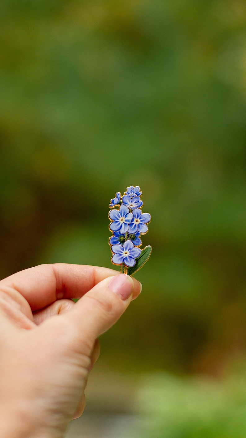 May include: A gold-toned enamel pin featuring a cluster of blue forget-me-not flowers with green leaves.