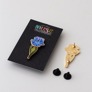 May include: A gold-toned enamel pin featuring a blue iris flower with green leaves. The pin is on a black card with the text "The Gray Muse" in white. Two black rubber pin backs are also included.