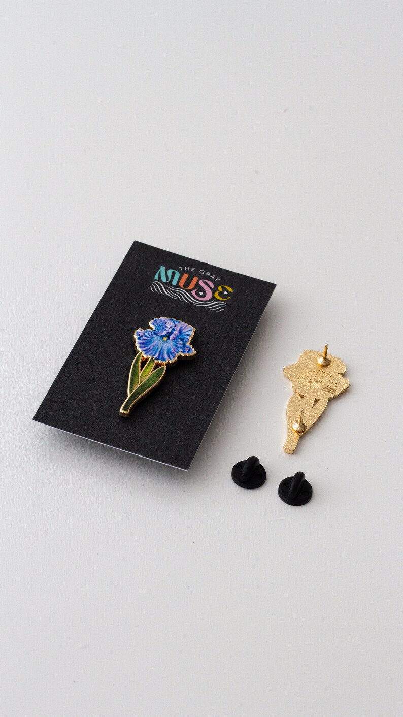 May include: A gold-toned enamel pin featuring a blue iris flower with green leaves. The pin is on a black card with the text "The Gray Muse" in white. Two black rubber pin backs are also included.
