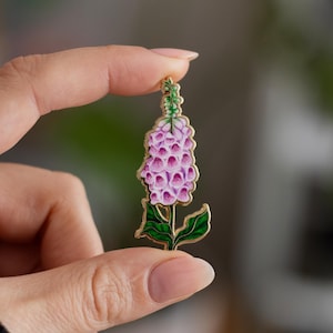 May include: A gold-tone enamel pin featuring a pink and green foxglove flower.