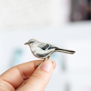 May include: A detailed enamel pin depicting a Northern Mockingbird. The bird is primarily white and gray with black wing markings and a gold-colored metal outline. The pin is held in a hand against a blurred background.