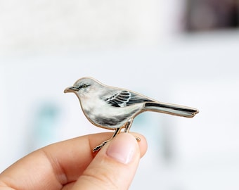 Puede incluir: Un broche de esmalte metálico con forma de pájaro pequeño con plumaje gris, blanco y negro. El ave se muestra de perfil mirando hacia la izquierda, con un contorno de metal dorado y detalles marcados en las plumas de sus alas y cola.