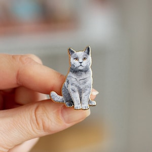Puede incluir: Un pin de esmalte de un gato gris con detalles dorados. El gato está sentado con la cabeza ligeramente inclinada hacia un lado y los ojos mirando directamente al espectador.