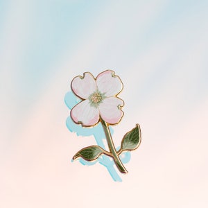 K&ouml;nnte beinhalten: Blumenbrosche mit einer zarten rosa-wei&szlig;en Blume, goldfarbenen Konturen und Stiel. Die Brosche hat zwei gr&uuml;ne Bl&auml;tter und ist vor einem weichen, farbverlaufenden Hintergrund platziert.