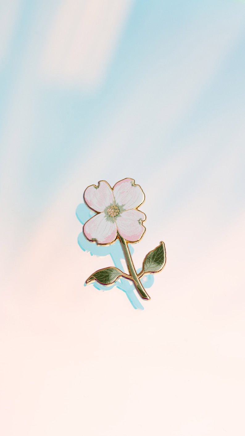 K&ouml;nnte beinhalten: Blumenbrosche mit einer zarten rosa-wei&szlig;en Blume, goldfarbenen Konturen und Stiel. Die Brosche hat zwei gr&uuml;ne Bl&auml;tter und ist vor einem weichen, farbverlaufenden Hintergrund platziert.
