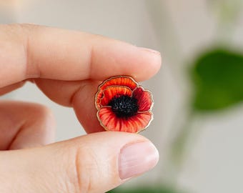 May include: A small enamel pin shaped like a poppy flower with vibrant red petals, a dark black center, and a thin gold-toned metal outline. The design features realistic shading on the petals, capturing the delicate texture of the flower.