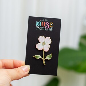 K&ouml;nnte beinhalten: Eine bl&uuml;tenf&ouml;rmige Emaille-Anstecknadel mit goldenem Umriss, hellrosa Bl&uuml;tenbl&auml;ttern und gr&uuml;nen Bl&auml;ttern. Die Anstecknadel ist an einer schwarzen Karte mit dem Text "THE GRAY MUSE" in bunten Buchstaben befestigt. Die Anstecknadel wird von einer Hand gehalten.