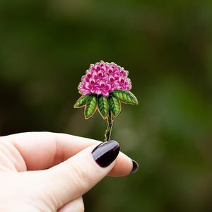 May include: A pink and green floral brooch. The brooch features a cluster of pink flowers with green leaves and a gold-colored stem. The brooch is held between two fingers against a blurred green background.